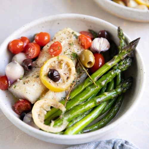 Two plated serving bowls with roasted asparagus and lemon baked cod topped with olives, cherry tomatoes, and fresh thyme.