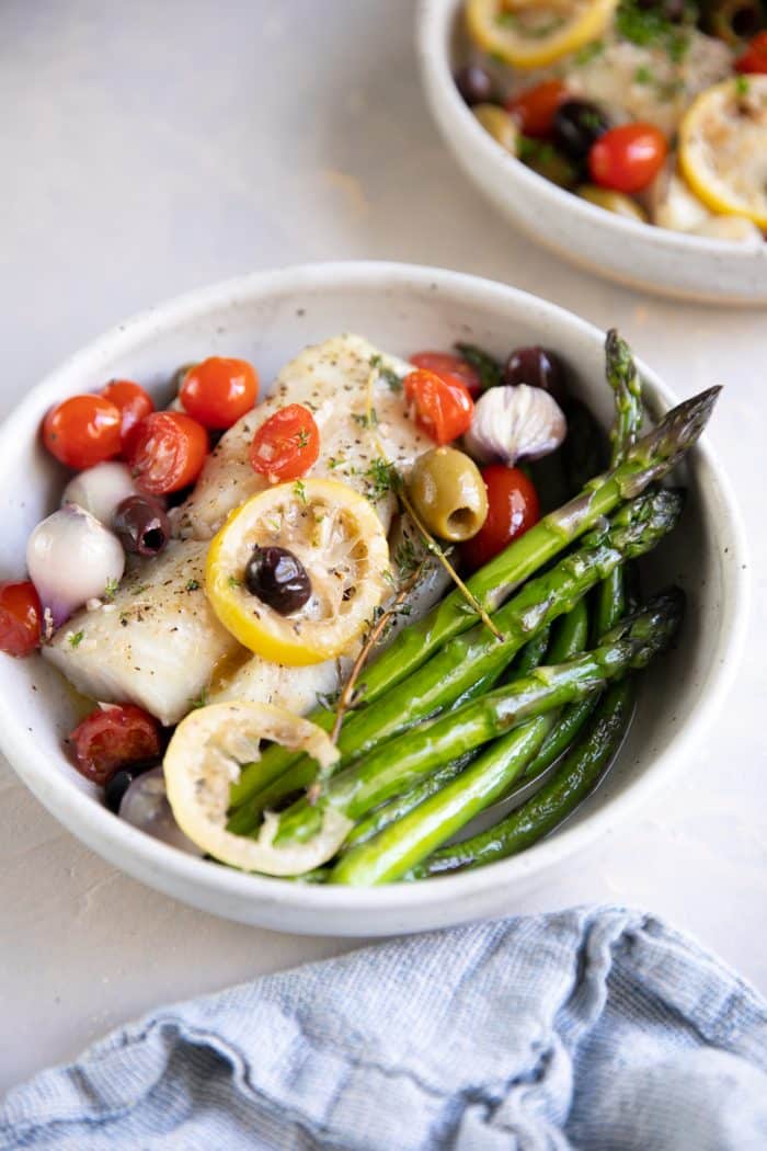 Two plated serving bowls with roasted asparagus and lemon baked cod topped with olives, cherry tomatoes, and fresh thyme.