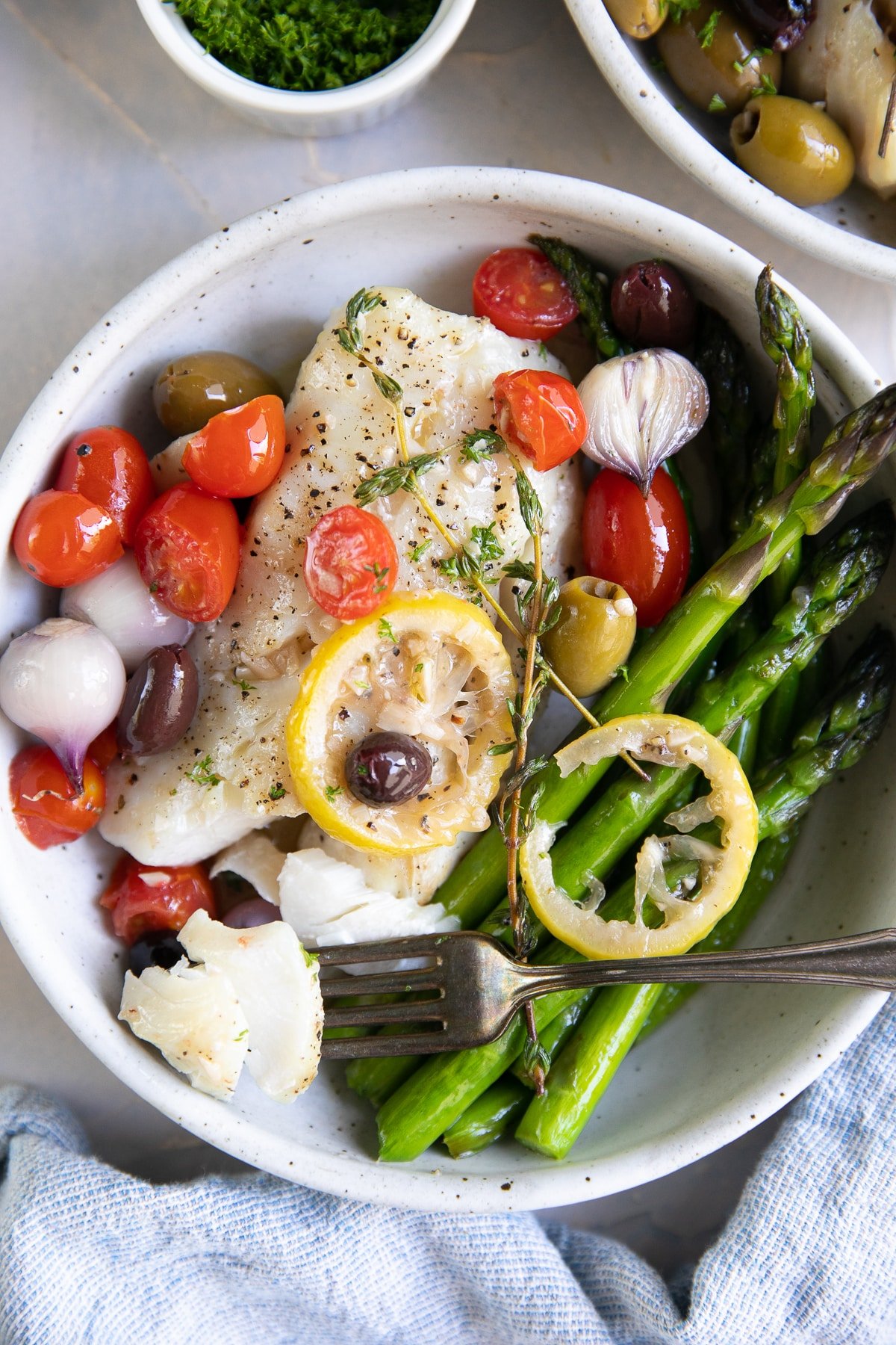 Two plated serving bowls with roasted asparagus and lemon baked cod topped with olives, cherry tomatoes, and fresh thyme.