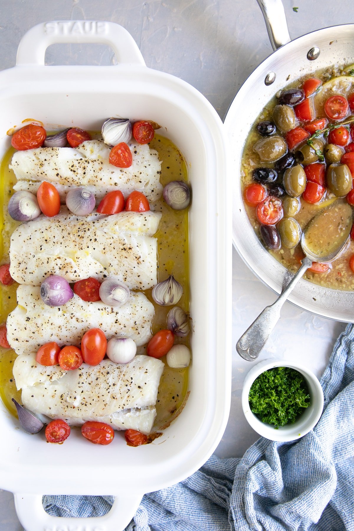 White baking dish filled with four cod fillets, cherry tomatoes, and pearl onions in a lemon butter sauce next to a skillet filled with lemon, garlic, and butter sauce and olives.