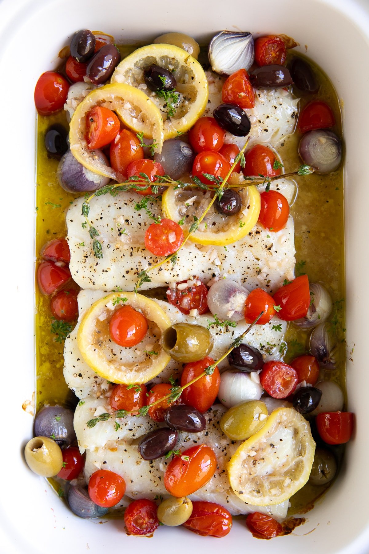 White baking dish filled with four cod fillets, cherry tomatoes, and pearl onions topped with cooked olives and cherry tomatoes in a lemon garlic white wine sauce.