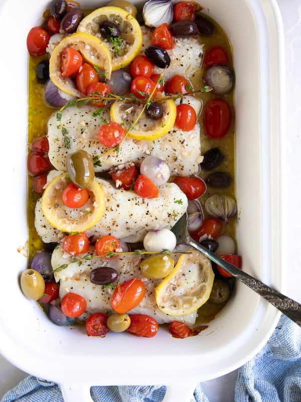 White baking dish filled with four cod fillets, cherry tomatoes, and pearl onions topped with cooked olives and cherry tomatoes in a lemon garlic white wine sauce.