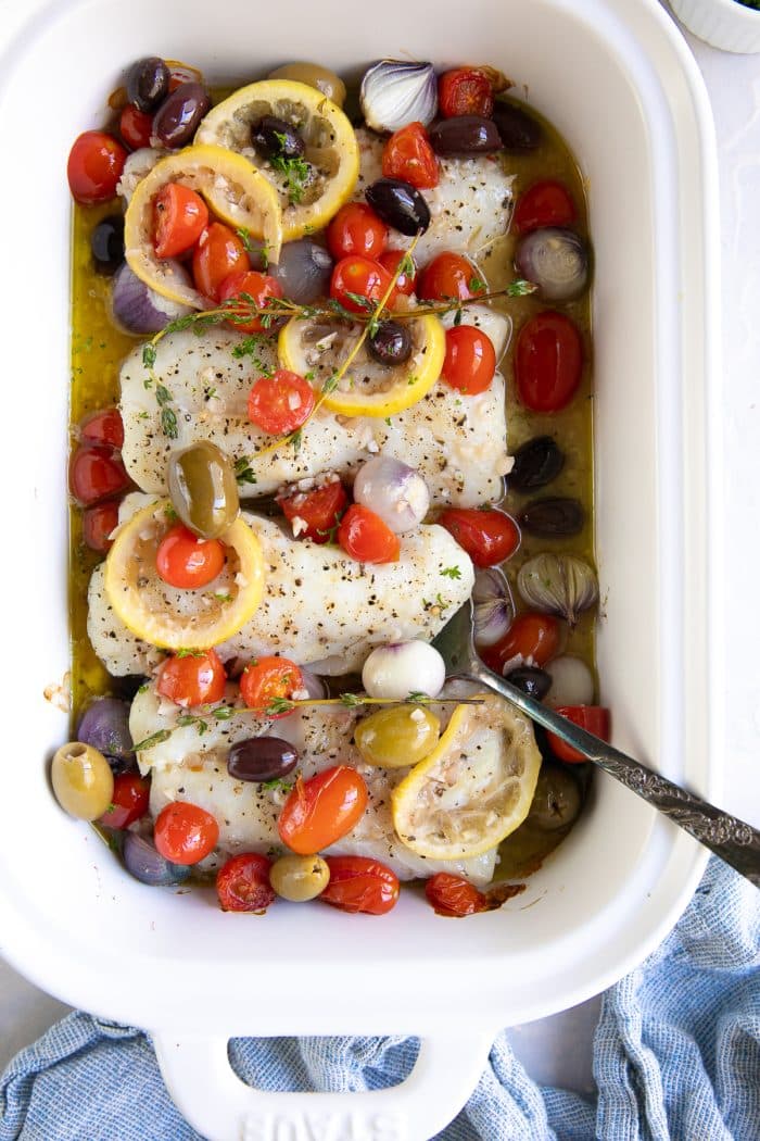 White baking dish filled with four cod fillets, cherry tomatoes, and pearl onions topped with cooked olives and cherry tomatoes in a lemon garlic white wine sauce.