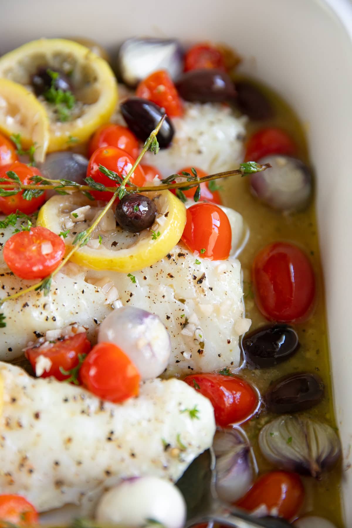 White baking dish filled with four cod fillets, cherry tomatoes, and pearl onions topped with cooked olives and cherry tomatoes in a lemon garlic white wine sauce.