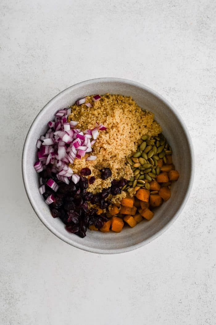 White mixing bowl filled with cooked white quinoa, pumpkin seeds, cooked butternut squash, dried cranberries, and diced red onion in individual sections.