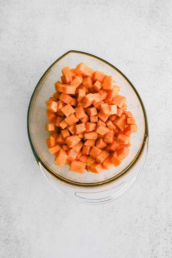 Glass mixing bowl filled with cubes of butternut squash tossed in oil and seasoned with salt and black pepper.