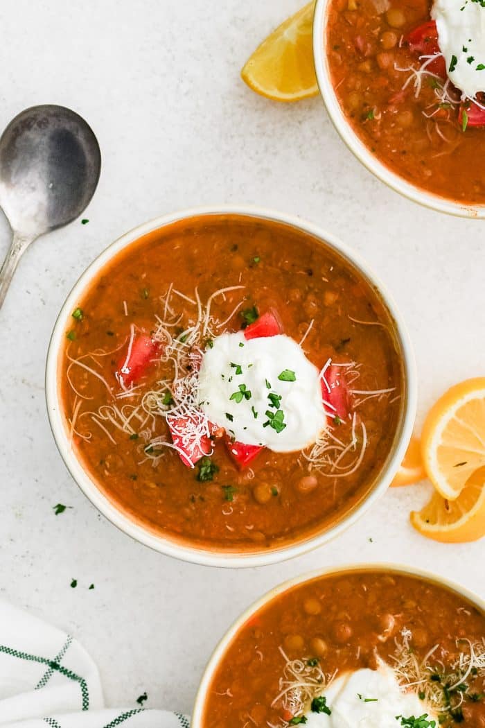 White bowl filled with lentil soup and topped with diced red tomato, shredded parmesan cheese, cream cheese, and chopped parsley.