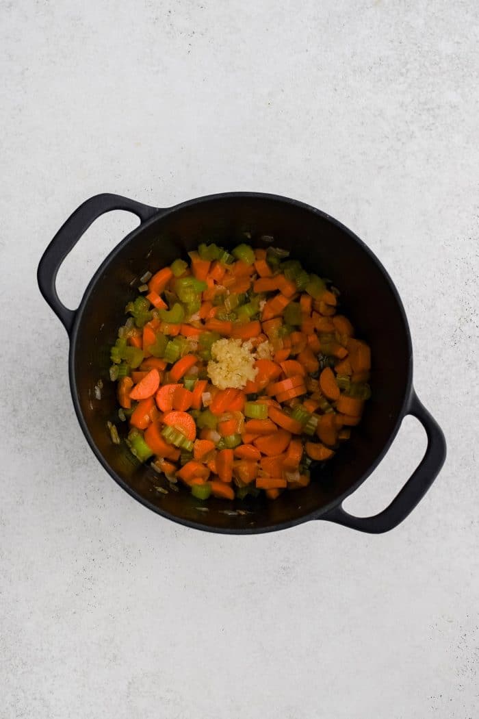 Minced garlic added to softened onion, carrots, and celery cooking in a large cast iron pot.