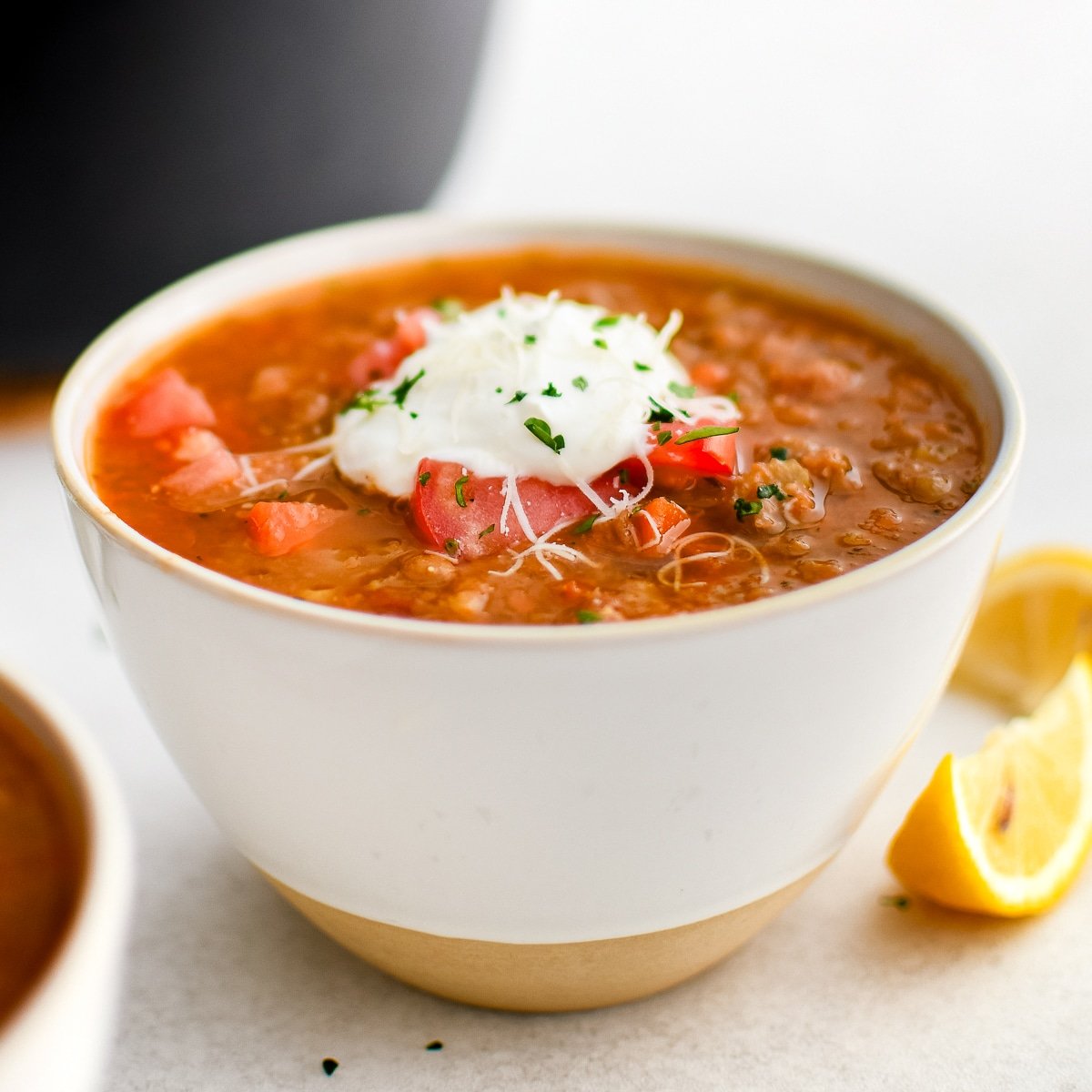 White bowl filled with lentil soup and topped with diced red tomato, shredded parmesan cheese, cream cheese, and chopped parsley.