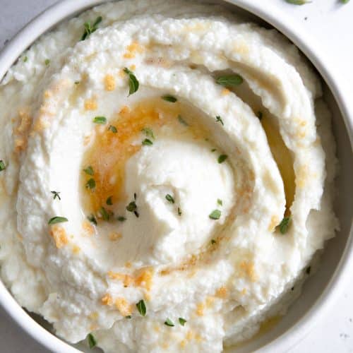 White serving bowl filled with creamy mashed cauliflower drizzled with brown butter and garlic.