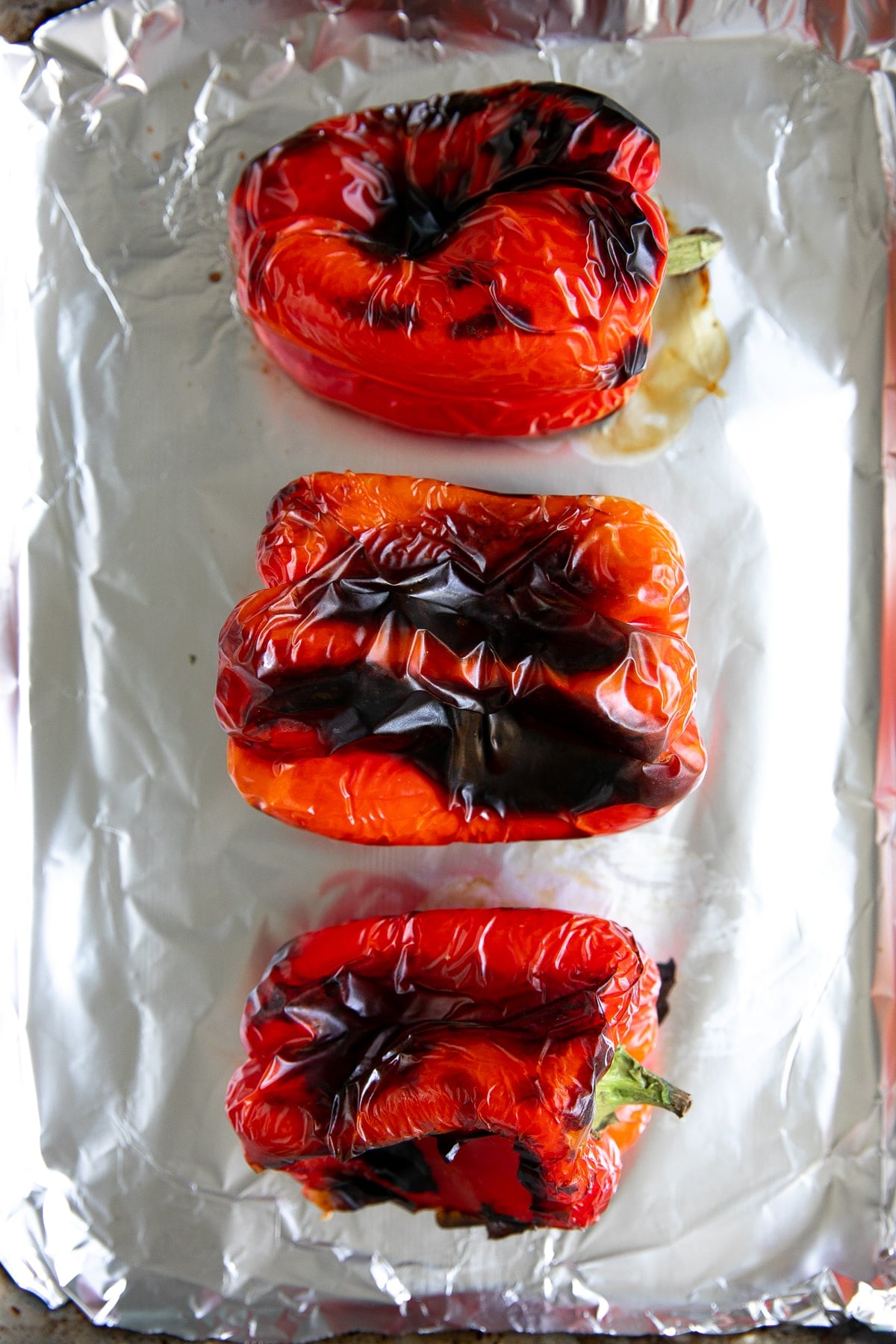 Three whole roasted red bell peppers with blackened skin on a large baking sheet.