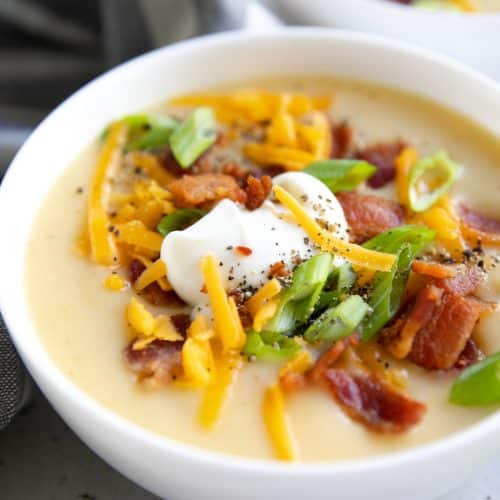 White bowl filled with creamy potato soup and topped with green onions, sour cream, cheddar cheese, and bacon.