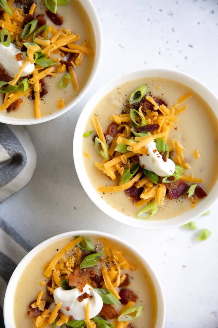 Three white bowls filled with creamy potato soup and topped with green onions, sour cream, cheddar cheese, and bacon.