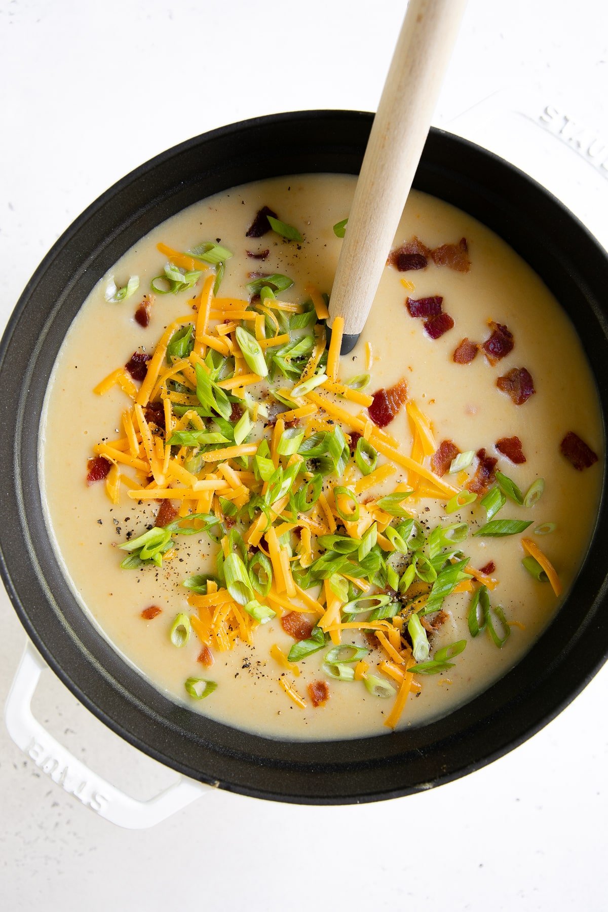 Large white soup pot filled with blended potato soup topped with chopped bacon, shredded cheddar cheese, green onions, and sour cream.
