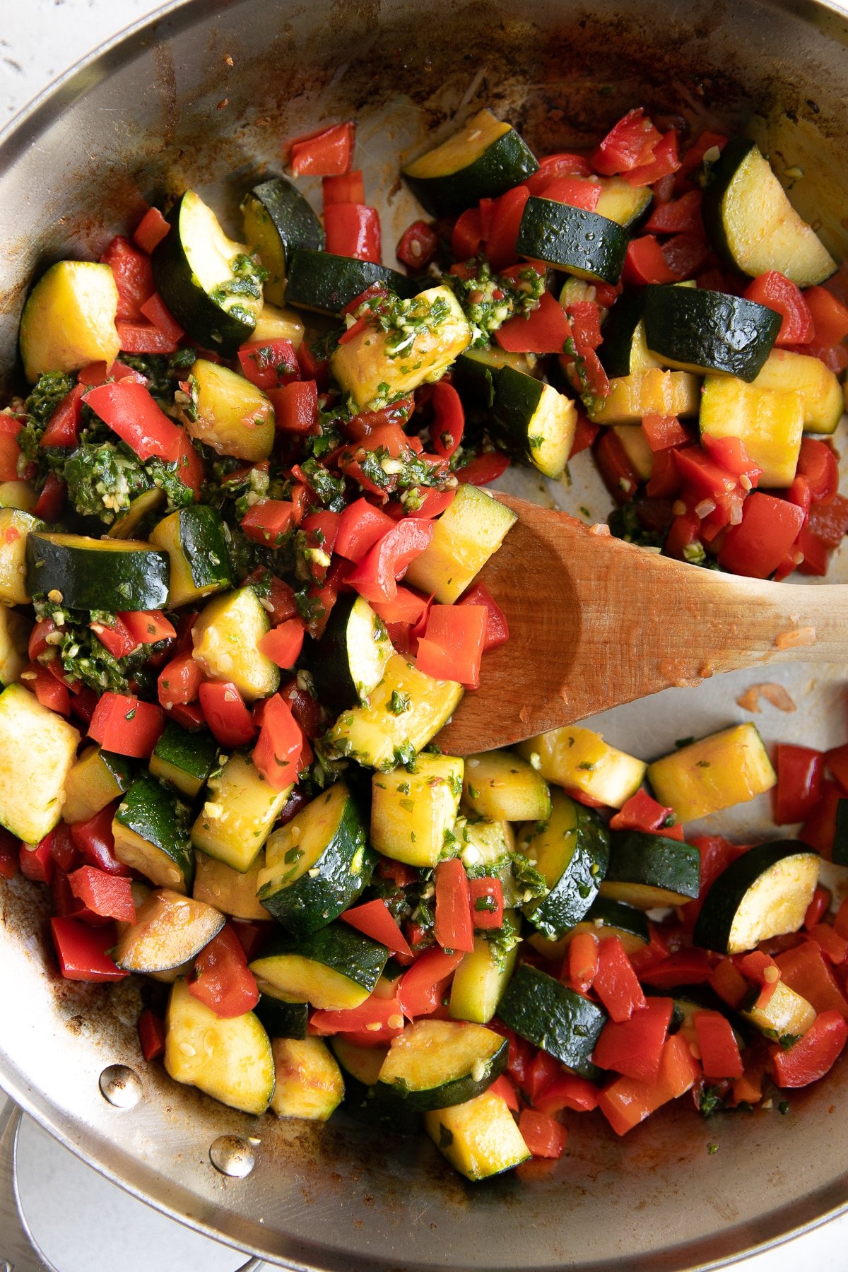 Zucchini and bell peppers mixing with homemade pestata in a large stainless steel skillet.