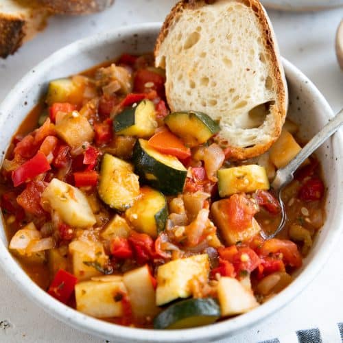 White shallow bowl filled with homemade Italian ratatouille and served with fresh toasted bread.