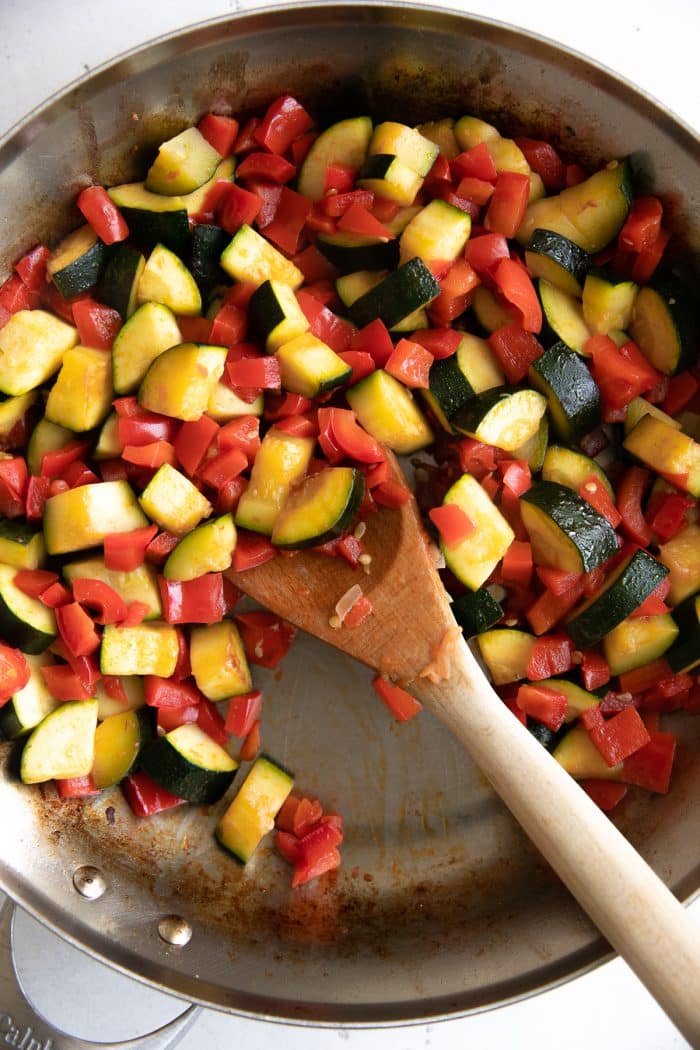 Sauteing zucchini, yellow squash, and chopped bell peppers in a large stainless steel skillet.