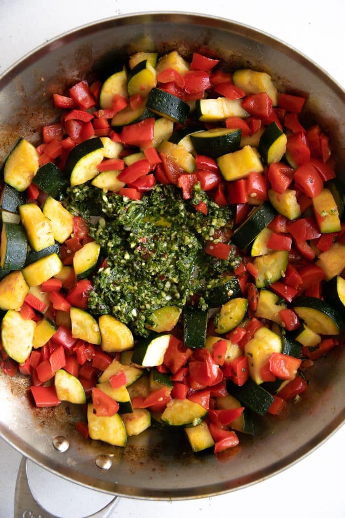 Zucchini and bell peppers mixing with homemade pestata in a large stainless steel skillet.