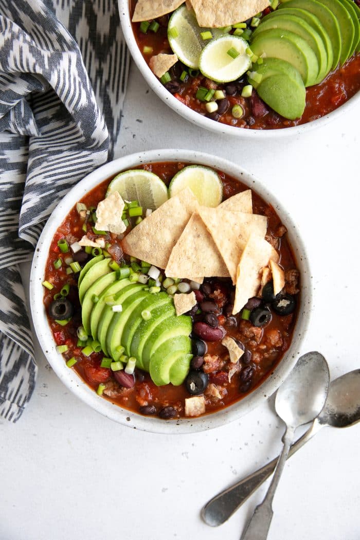 Overhead image of two white shallow bowls filled with homemade taco soup and topped with sliced avocado, tortilla chips, green onions, and lime wedges.