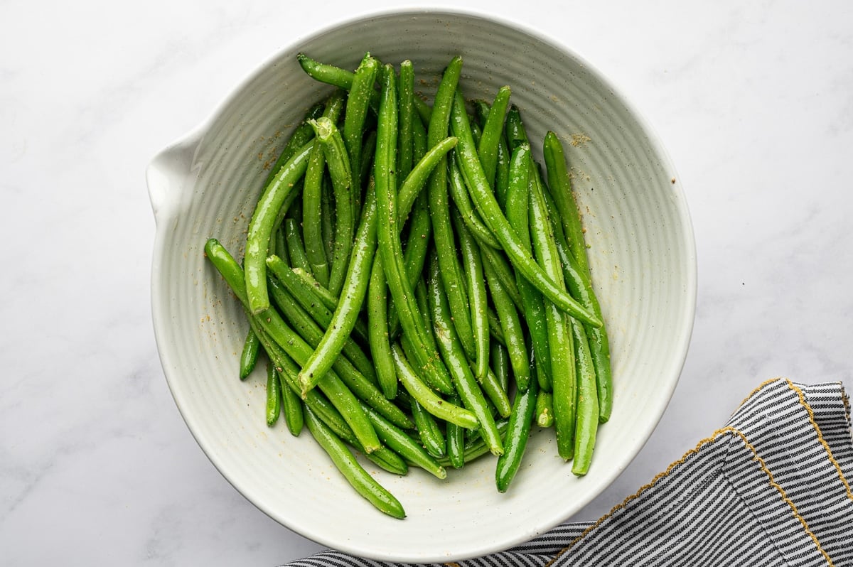Green beans coated in olive oil and seasonings in a white bowl