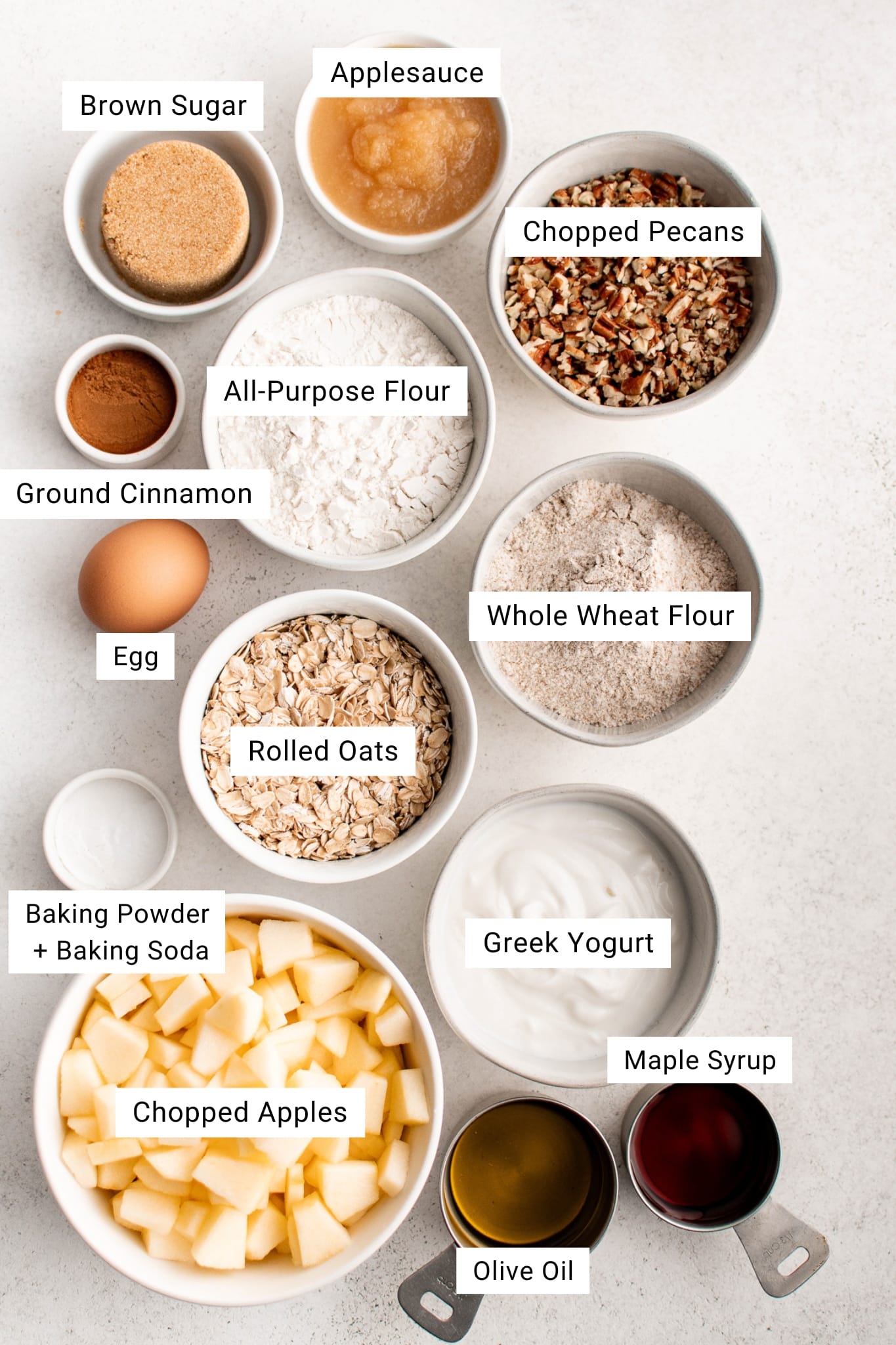 Overhead image of individual apple cinnamon bread ingredients arranged in small bowls and labeled, including brown sugar, applesauce, chopped pecans, ground cinnamon, all-purpose flour, whole wheat flour, rolled oats, Greek yogurt, chopped apples, olive oil, maple syrup, an egg, and a small bowl with baking powder and baking soda.