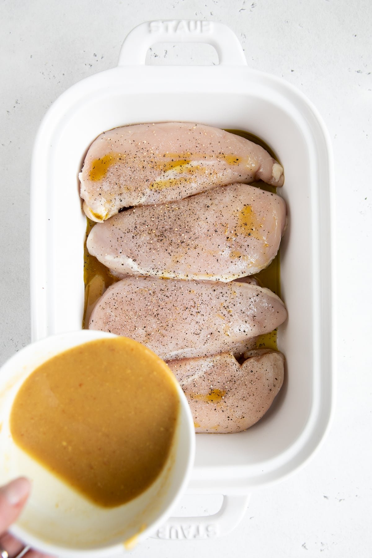 Four raw chicken breasts seasoned with salt and pepper in a white baking dish.