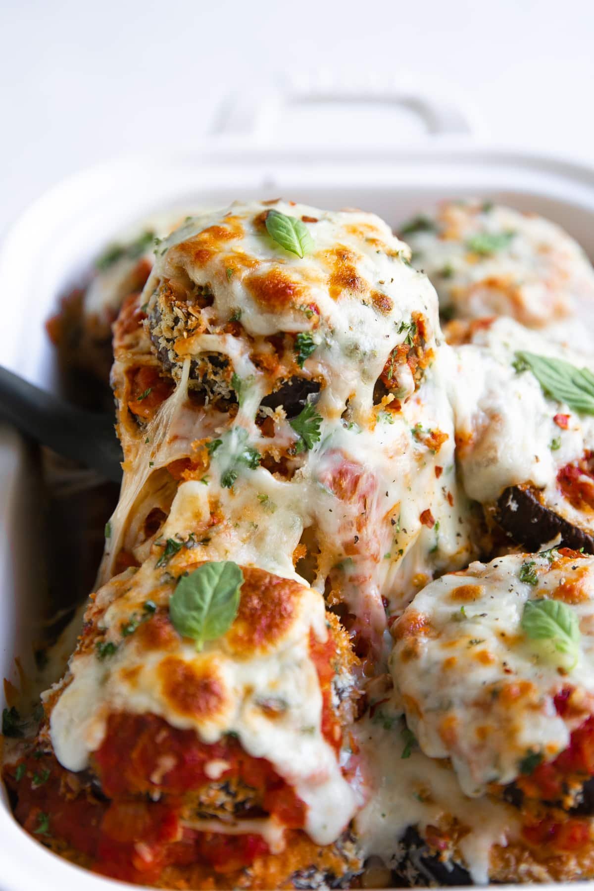 Spatula removing one eggplant parmesan stack from a white baking dish.