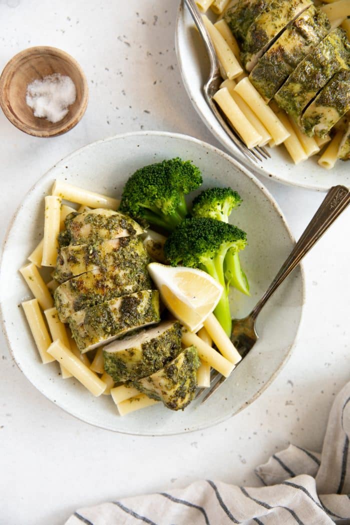 Baked pesto chicken recipe on a white serving plate with cooked pasta noodles, fresh lemon wedges, and steamed broccoli.