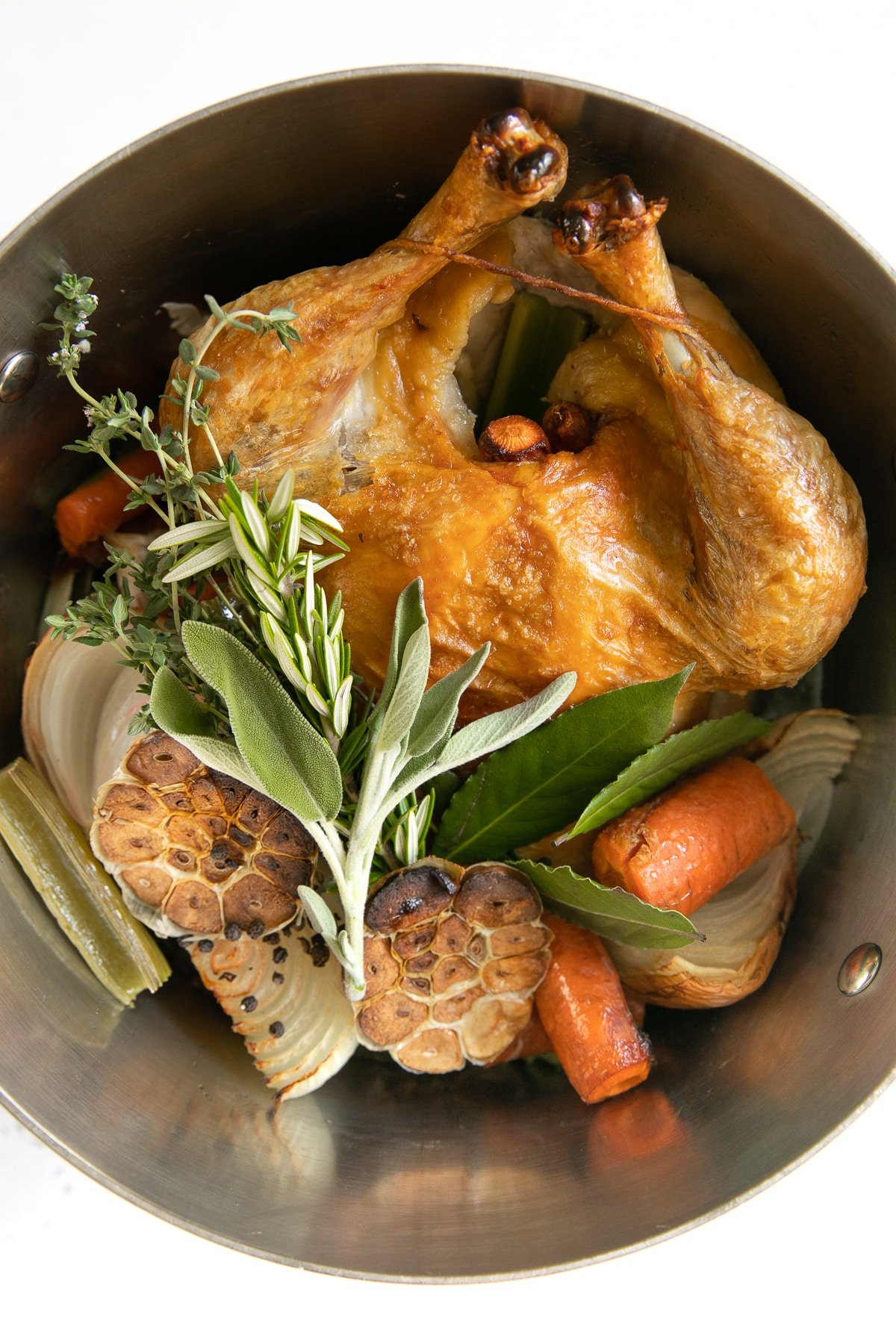 Large stock pot filled with one whole browned chicken, garlic, vegetables, and fresh herbs.