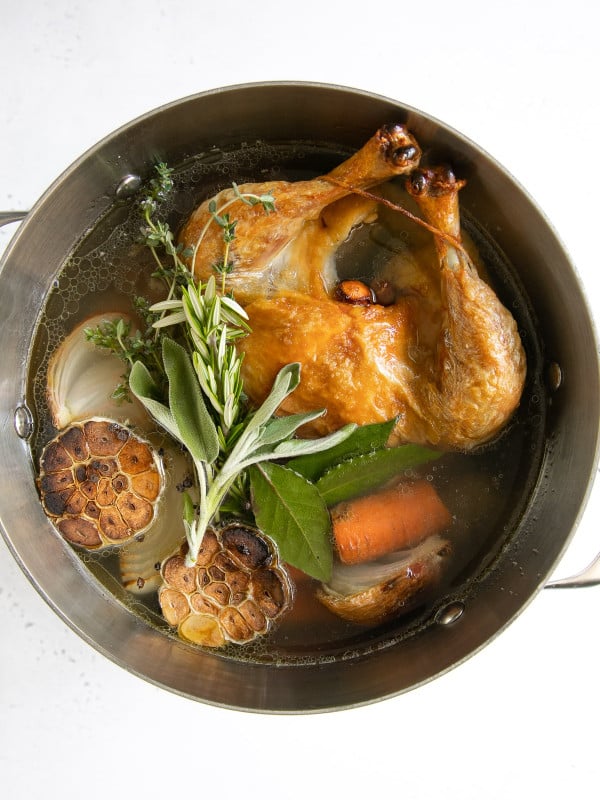 Large stock pot filled with one whole browned chicken, garlic, vegetables, and fresh herbs.