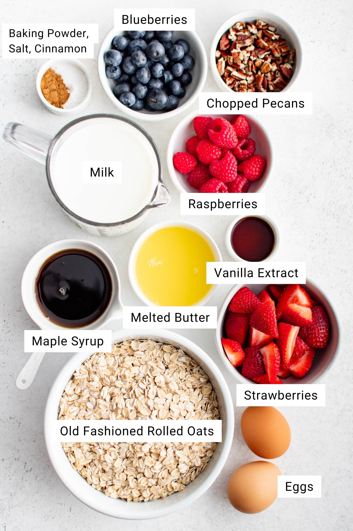 Overhead view of labeled ingredients for baked oatmeal including oats, milk, eggs, berries, pecans, butter, maple syrup, and cinnamon.