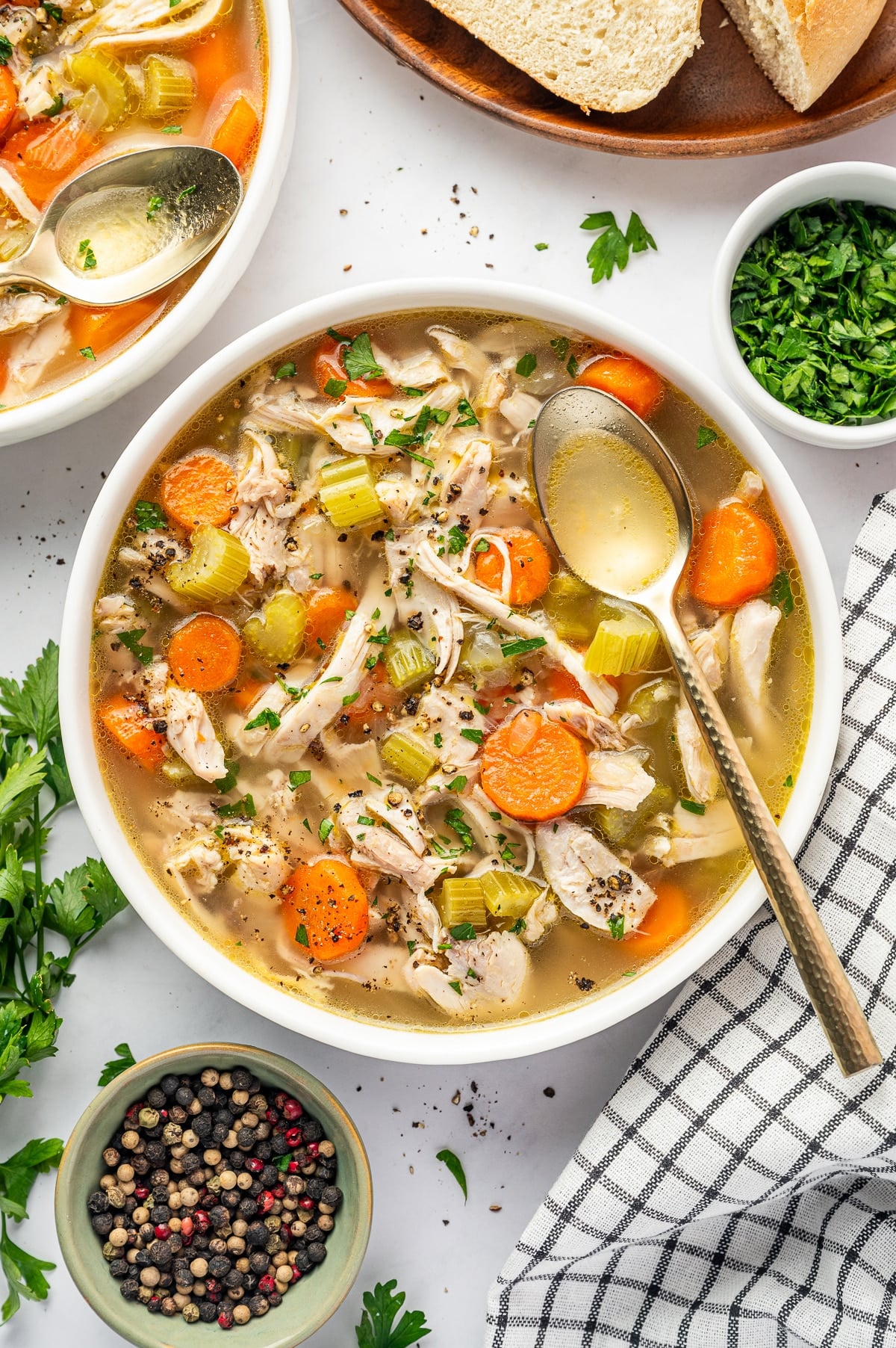 Overhead view of chicken soup served in a wide white bowl, filled with shredded chicken, sliced carrots, celery, onion, and clear golden broth, topped with parsley and black pepper and served with a metal spoon, bread, and fresh parsley.