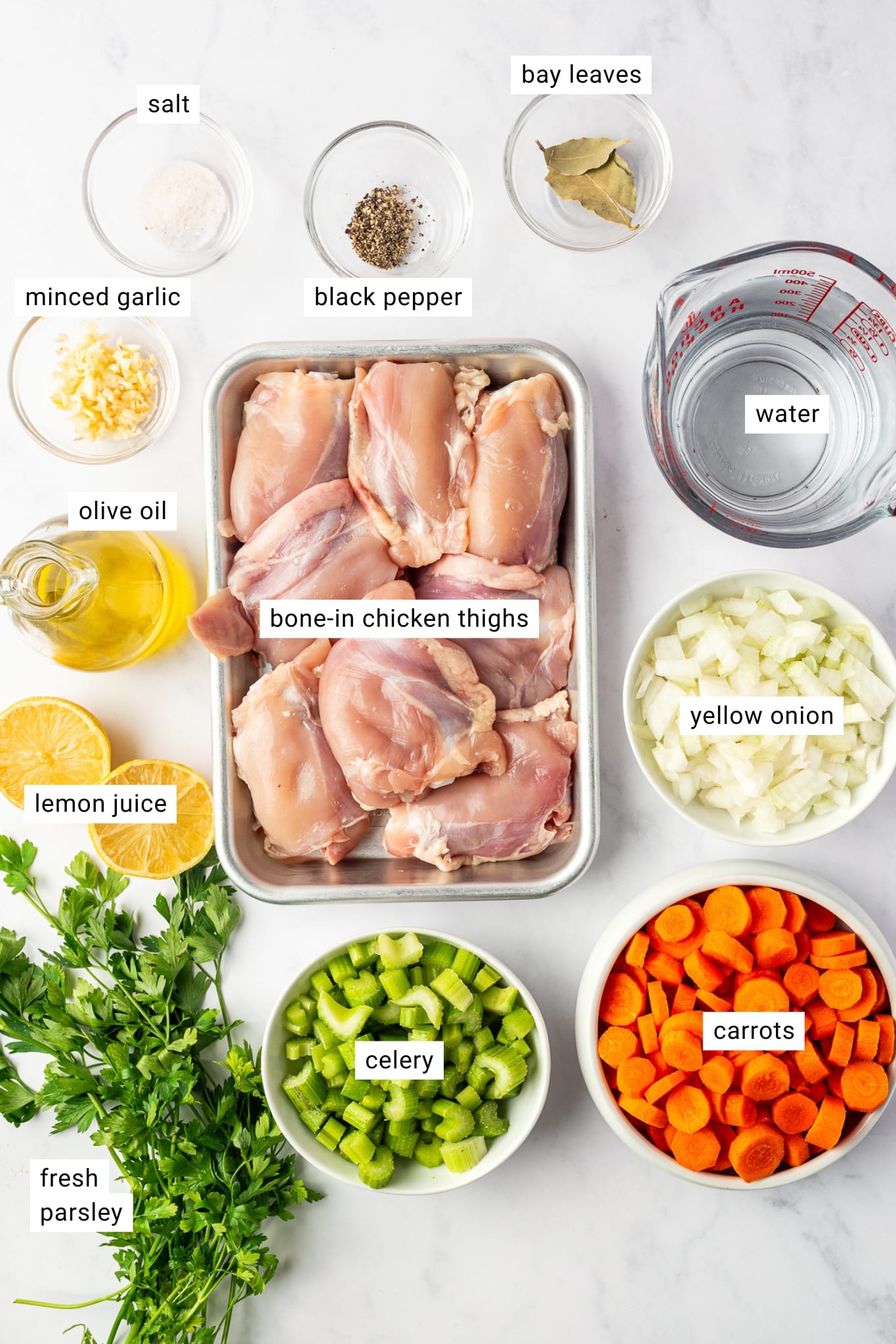 Overhead layout of chicken soup ingredients with text labels identifying salt, black pepper, bay leaves, water, minced garlic, olive oil, bone-in chicken thighs, yellow onion, lemon juice, celery, carrots, and fresh parsley on a white background.