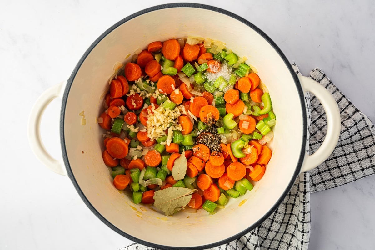 Overhead view of sauteed carrots, celery, and onion in a cream-colored Dutch oven topped with minced garlic, black pepper, salt, and two dried bay leaves before the broth is added.
