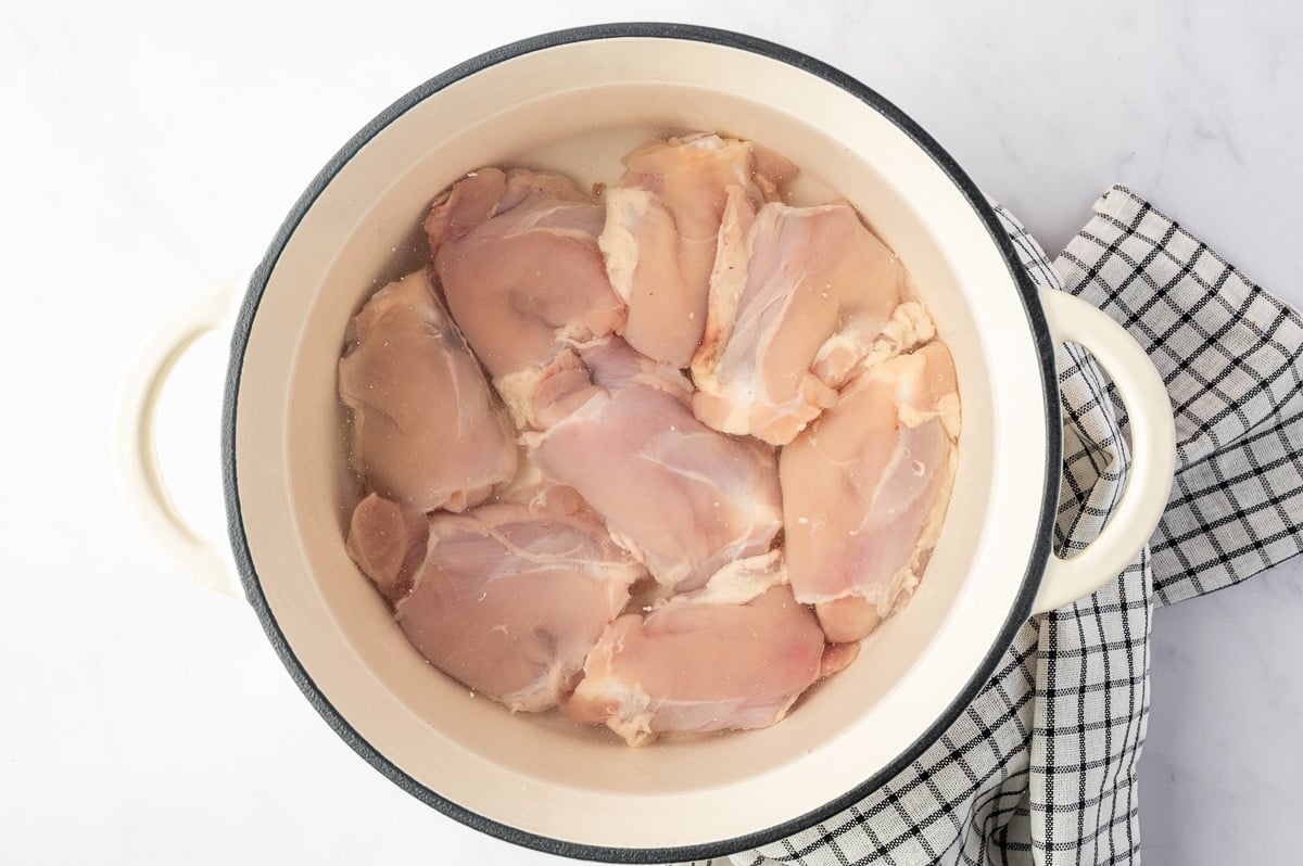 Overhead view of raw bone-in, skinless chicken thighs submerged in water inside a cream-colored enameled Dutch oven with a black rim, set beside a black-and-white checkered kitchen towel.