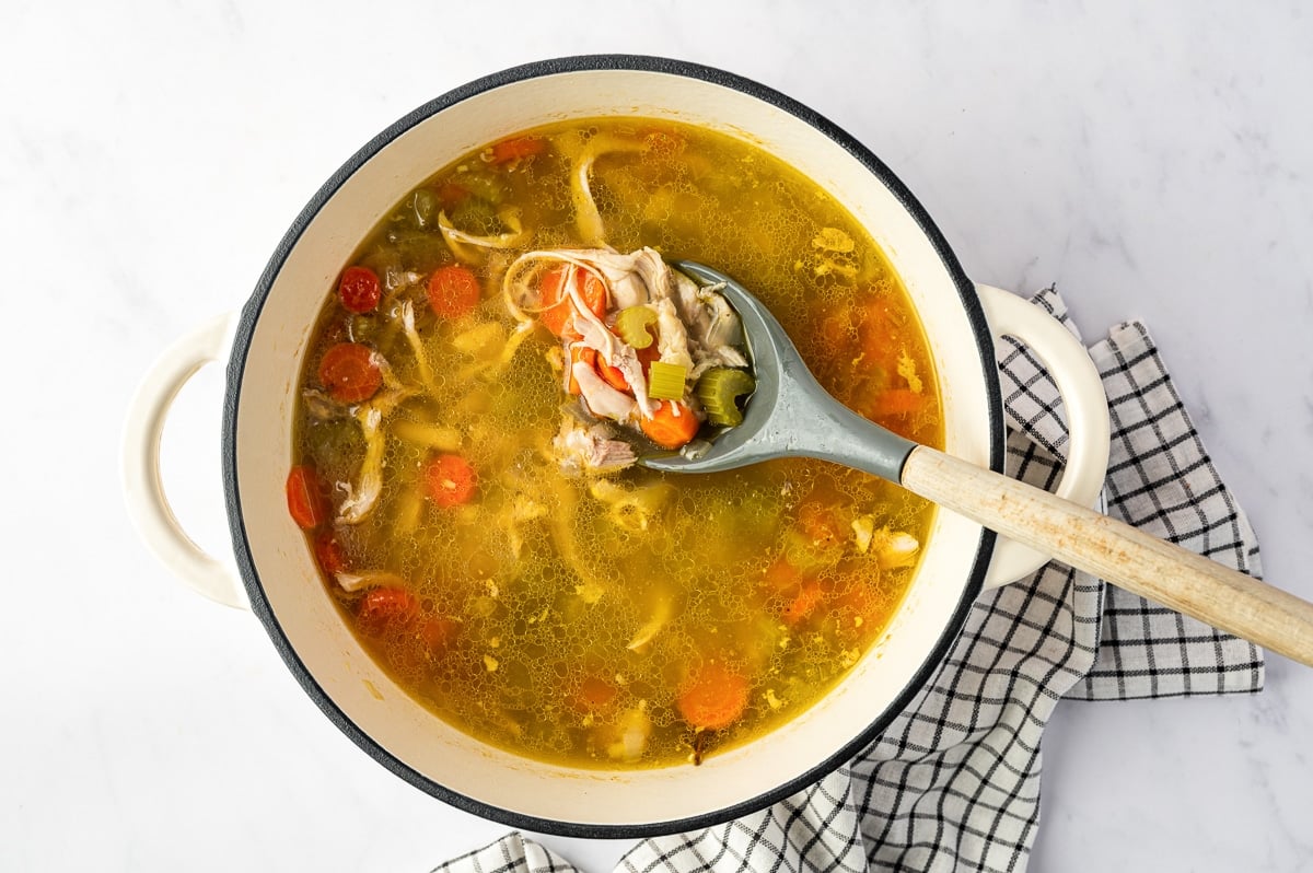 Overhead view of finished homemade chicken soup in a cream-colored enameled Dutch oven, filled with shredded chicken, sliced carrots, celery, and golden broth, with a gray ladle lifting a spoonful of chicken and vegetables.
