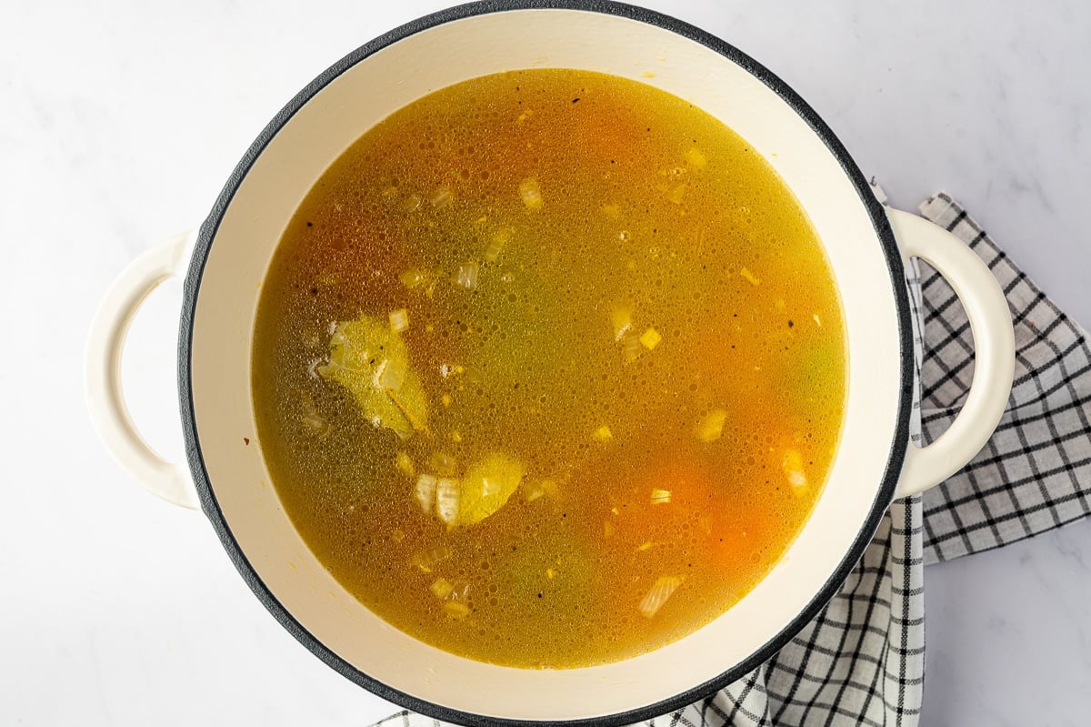 Overhead view of golden homemade chicken broth covering carrots, celery, onion, garlic, and bay leaves inside a cream-colored enameled Dutch oven with a black rim.