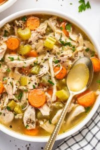 Angled close view of homemade chicken soup in a large white bowl filled with shredded chicken, orange carrot rounds, celery, onion, and golden broth, garnished with parsley and cracked black pepper with a metal spoon resting in the bowl.