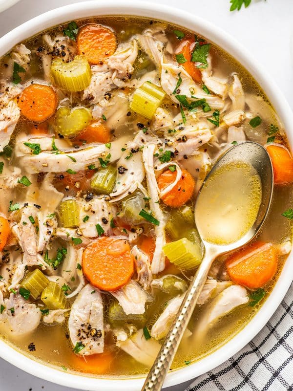Angled close view of homemade chicken soup in a large white bowl filled with shredded chicken, orange carrot rounds, celery, onion, and golden broth, garnished with parsley and cracked black pepper with a metal spoon resting in the bowl.