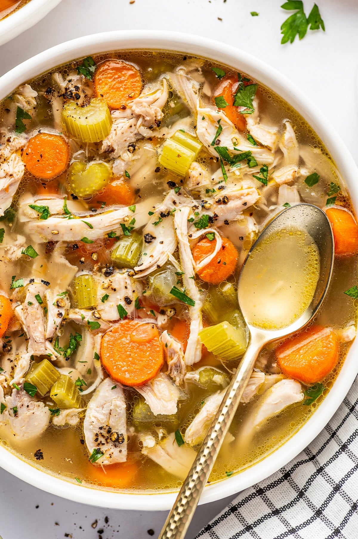 Angled close view of homemade chicken soup in a large white bowl filled with shredded chicken, orange carrot rounds, celery, onion, and golden broth, garnished with parsley and cracked black pepper with a metal spoon resting in the bowl.
