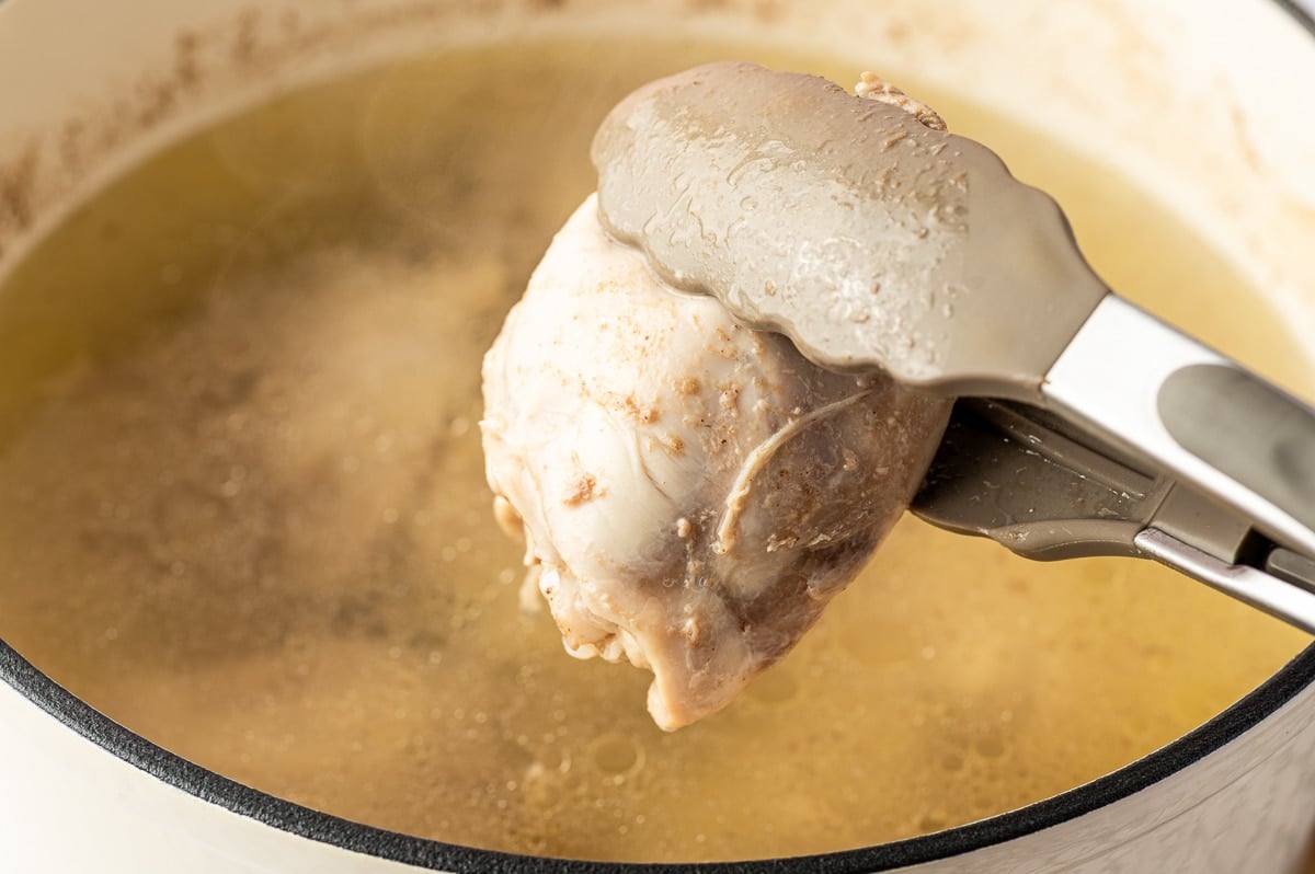 Close-up of metal kitchen tongs lifting a cooked chicken thigh from light golden broth in a cream-colored enameled Dutch oven after the chicken has finished simmering.