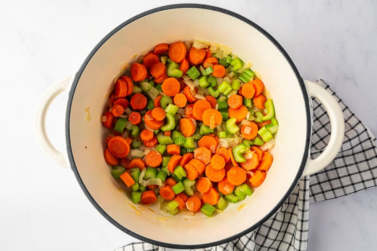 Overhead view of sliced orange carrots, chopped green celery, and diced yellow onion cooking together in a cream-colored enameled Dutch oven with a black rim.