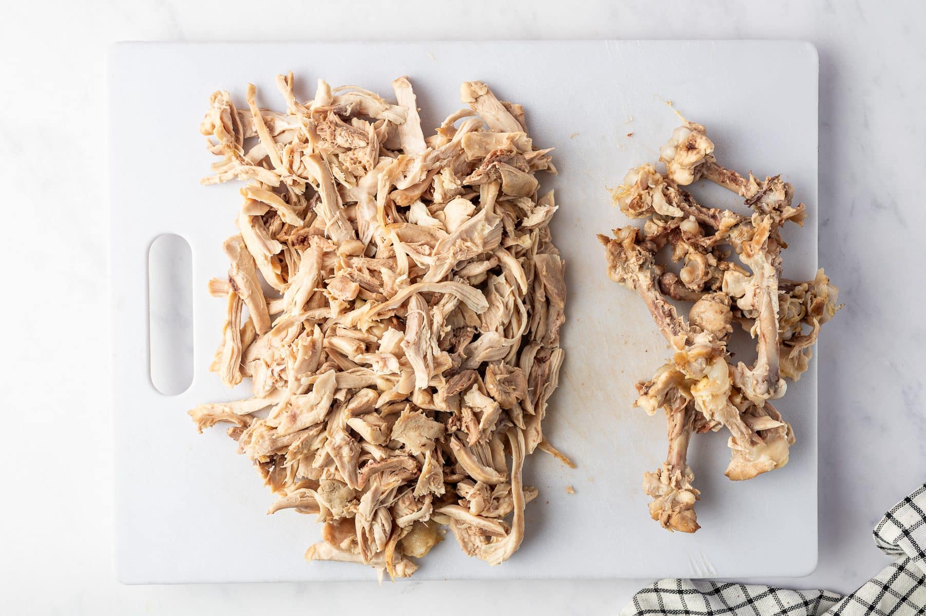 Overhead view of cooked chicken thigh meat shredded into bite-size pieces on a large white cutting board, with the discarded chicken bones separated at the bottom.
