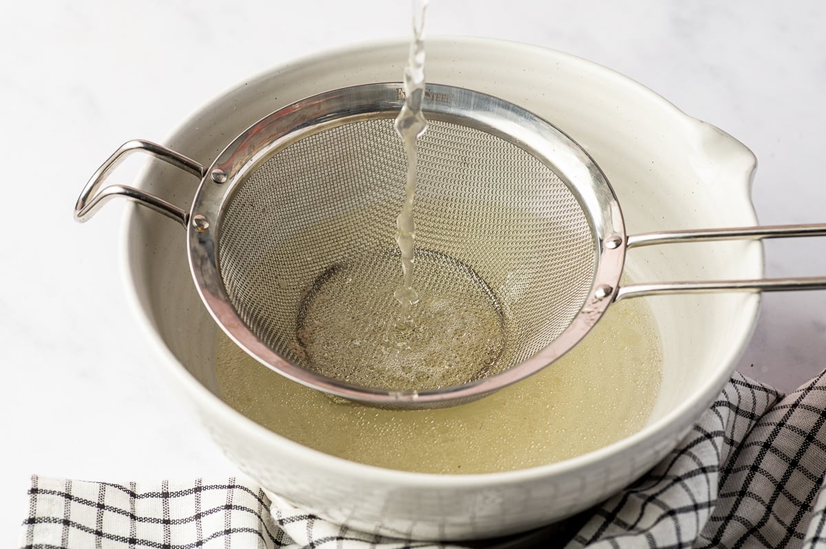 Light golden homemade chicken broth being poured through a fine-mesh metal strainer positioned over a large cream-colored ceramic bowl, with a black-and-white checkered towel alongside.