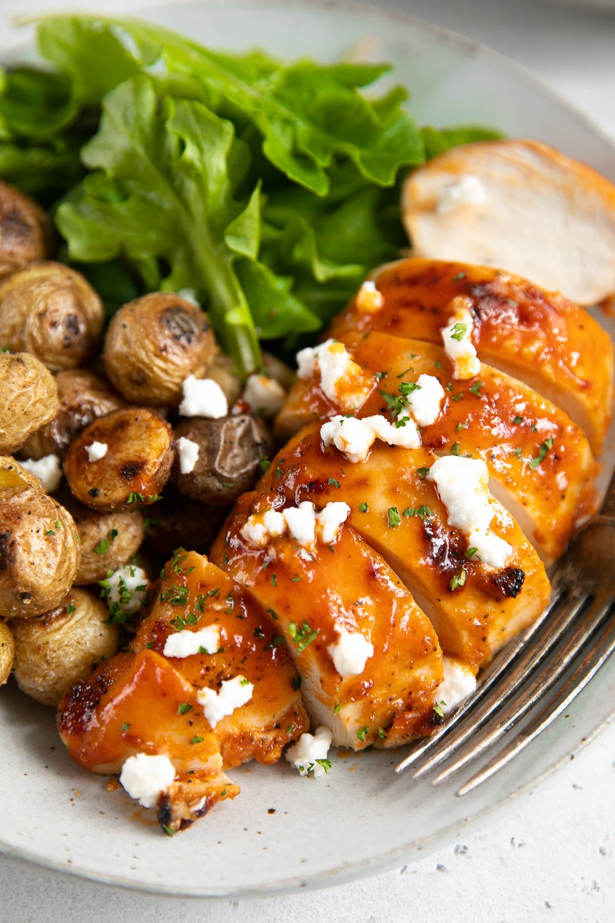 Baked buffalo chicken breast sliced and plated on a white serving plate and served with mini roasted potatoes and fresh greens.