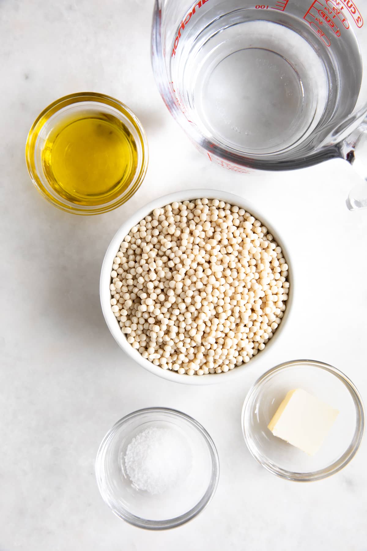 Individual bowls filled with olive oi, butter, salt, pearl couscous, and water.