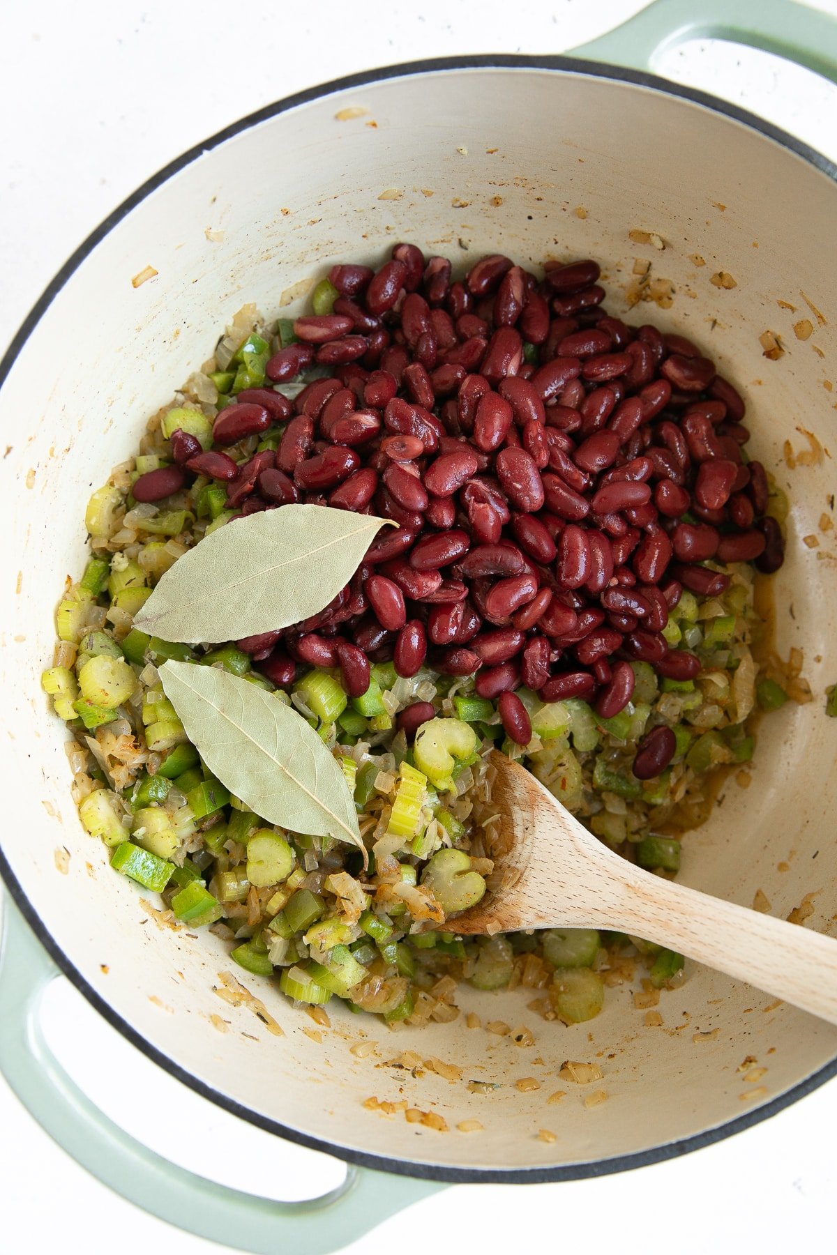 Large Dutch oven filled with cooked onions, celery, and green bell pepper with garlic and topped with red kidney beans and two bay leaves.