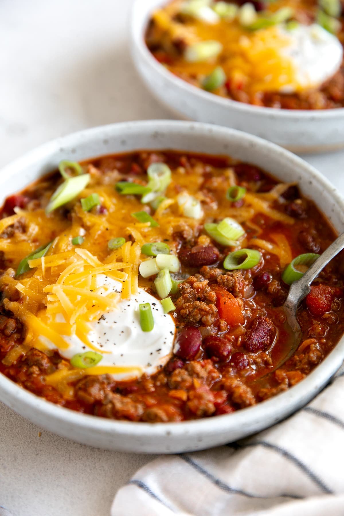 White shallow bowl filled with the best chili recipe and topped with shredded cheddar cheese, sour cream, and green onions.
