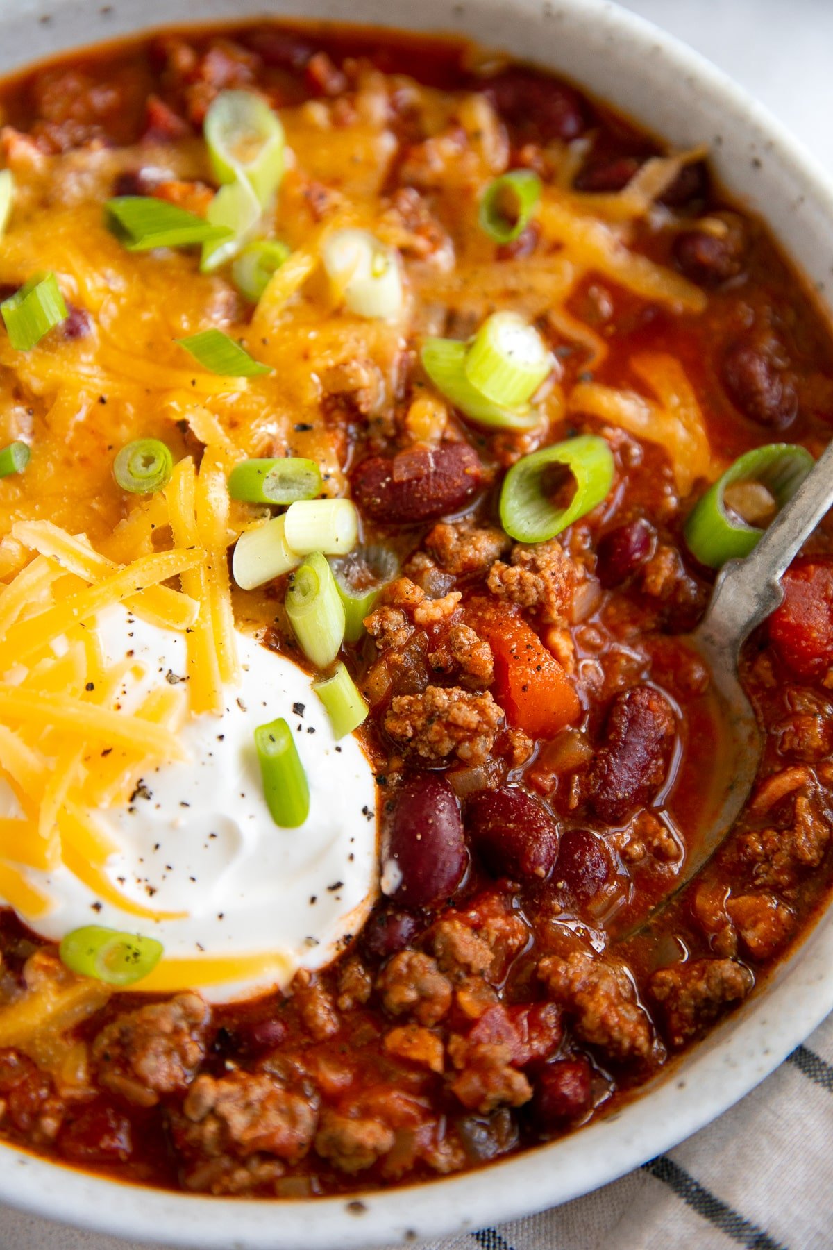 White shallow bowl filled with the best chili recipe and topped with shredded cheddar cheese, sour cream, and green onions.