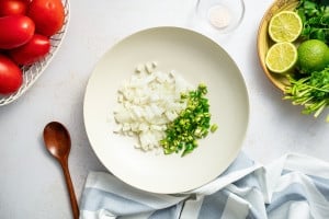 White diced onion and chopped green serrano peppers in a large mixing bowl, with fresh cilantro and halved limes nearby.